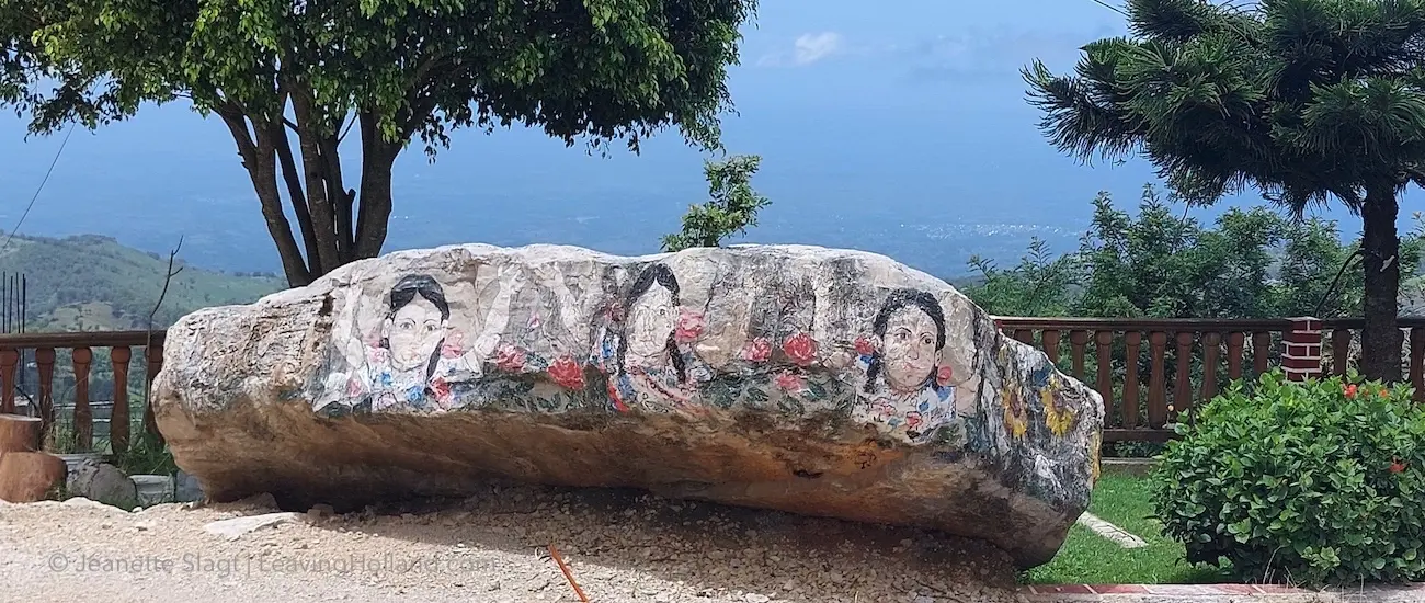 blauwe lucht grote platte steen op de rand van de tuin boven de vlakte beneden, met een schildering van Maya vrouwen in traditionele kleding op de zijkant, tussen elke vrouw een paar rozen