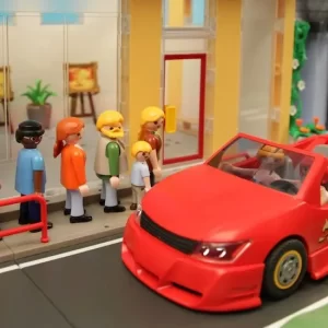palymobil poppetjes in een rij rond de hoek van een atelier, rode auto met juichend groen poppetje komt aanrijden.